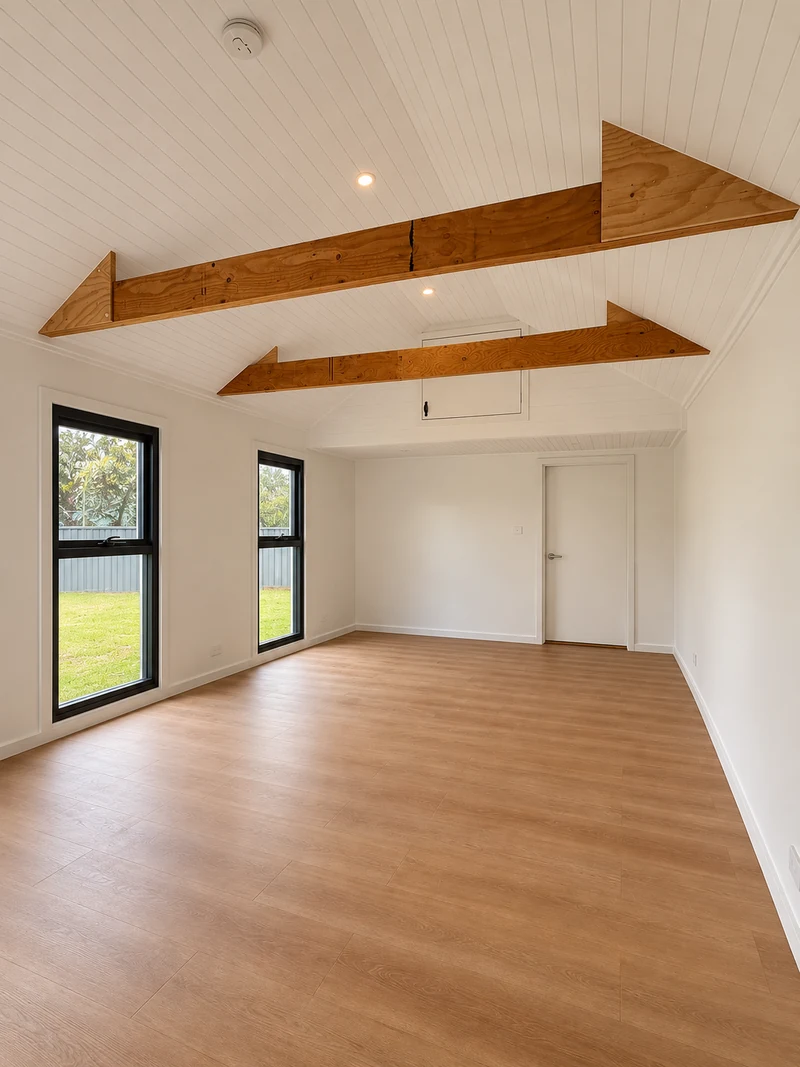 Bright converted room with exposed timber beams, a pitched white ceiling and garden-facing windows